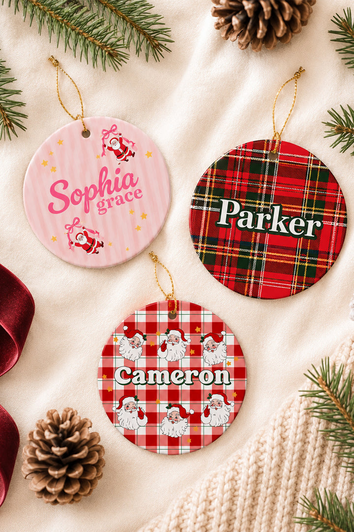 Three Caden Lane personalized round Christmas ornaments displayed on a cream fabric background with pine branches, pinecones, and a knit sweater. One ornament is pink with Santa graphics and the name Sophia Grace, one is red plaid with the name Parker, and one is red plaid with Santa graphics and the name Cameron.