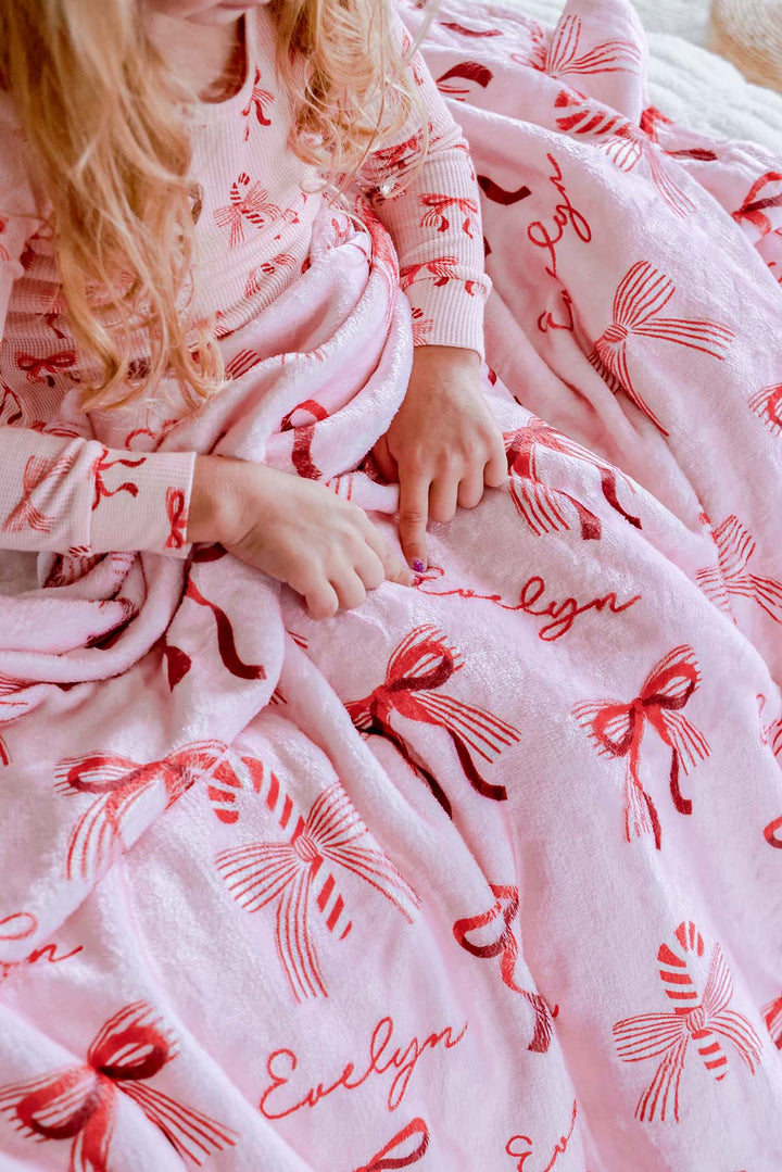 Child cuddled in the Caden Lane Personalized Peppermint Bows Kids' Christmas Blanket featuring festive red bows, candy canes, and a custom embroidered name.