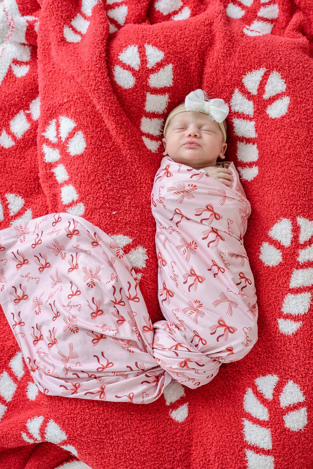 Baby sleeping in the Caden Lane Personalized Peppermint Bows Christmas Swaddle Blanket with matching personalized receiving blanket featuring festive holiday bows and candy canes.