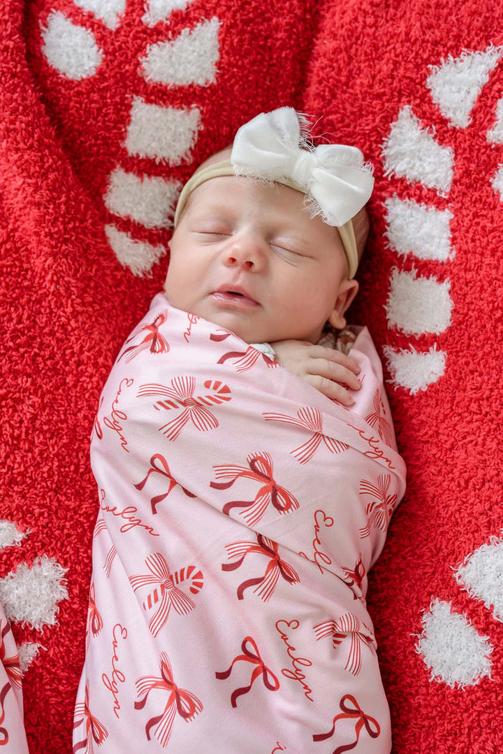 Newborn wrapped in the Caden Lane Personalized Peppermint Bows Christmas Swaddle Blanket featuring festive red bows, candy canes, and a custom name on a soft blush pink background.