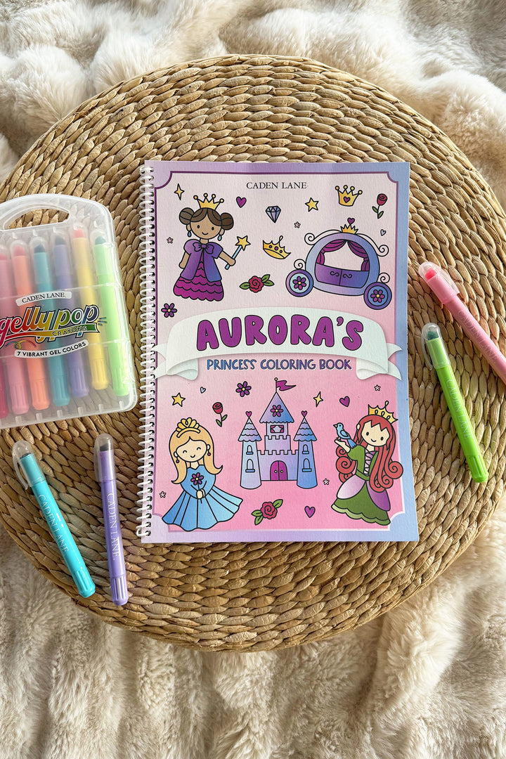 Caden Lane 'Aurora's Princess Coloring Book' on a woven tray with a cream throw, Gellypop gel crayon case, and pastel Caden Lane markers
