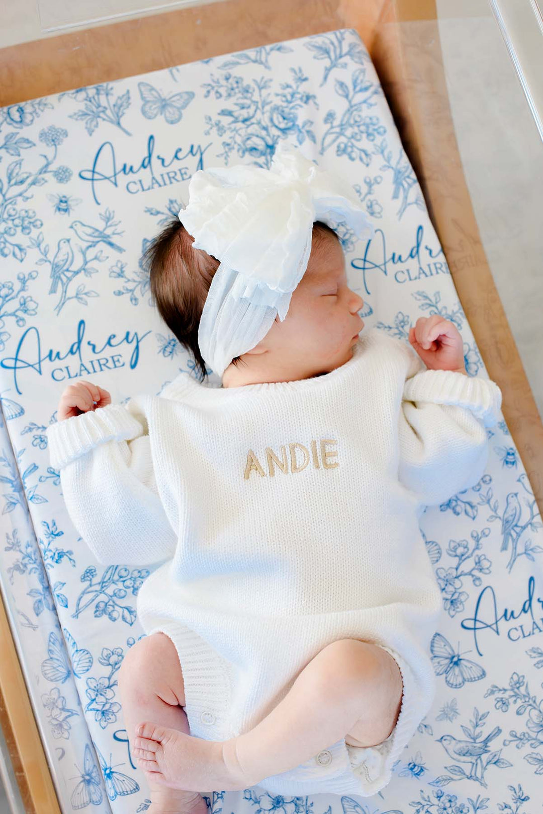 Full-length shot of a newborn, arms outstretched, on a Caden Lane blue knit swaddle in an Enchanted Meadow Toile print featuring florals, reading "Audrey Claire," paired with a white romper.
