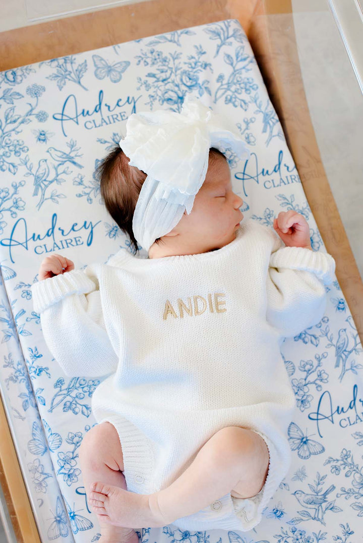Full-length shot of a newborn, arms outstretched, on a Caden Lane blue knit swaddle in an Enchanted Meadow Toile print featuring florals, reading "Audrey Claire," paired with a white romper.