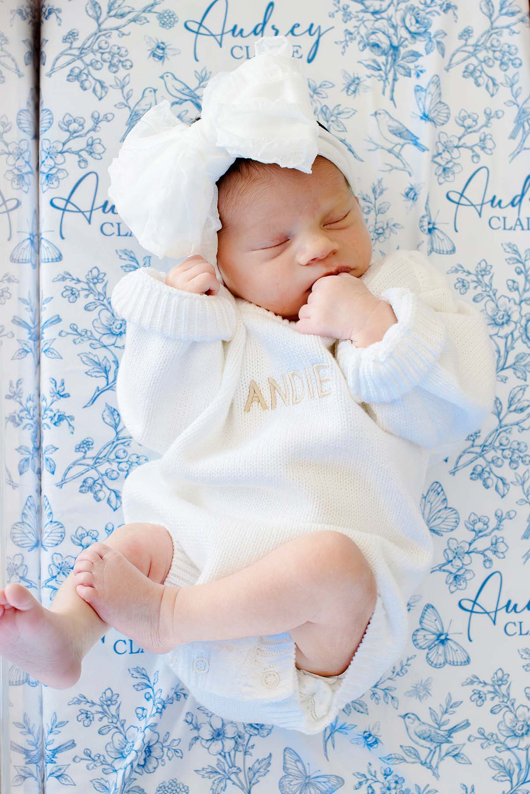 Overhead shot of a newborn, hand at mouth, on a Caden Lane blue knit swaddle in an Enchanted Meadow Toile print featuring butterflies, reading "Audrey Claire," paired with a romper.
