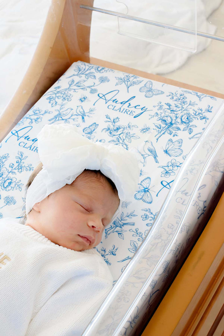 Close-up of a newborn asleep in a bassinet on a Caden Lane blue knit swaddle in an Enchanted Meadow Toile print featuring birds, reading "Audrey Claire," paired with a headband.
