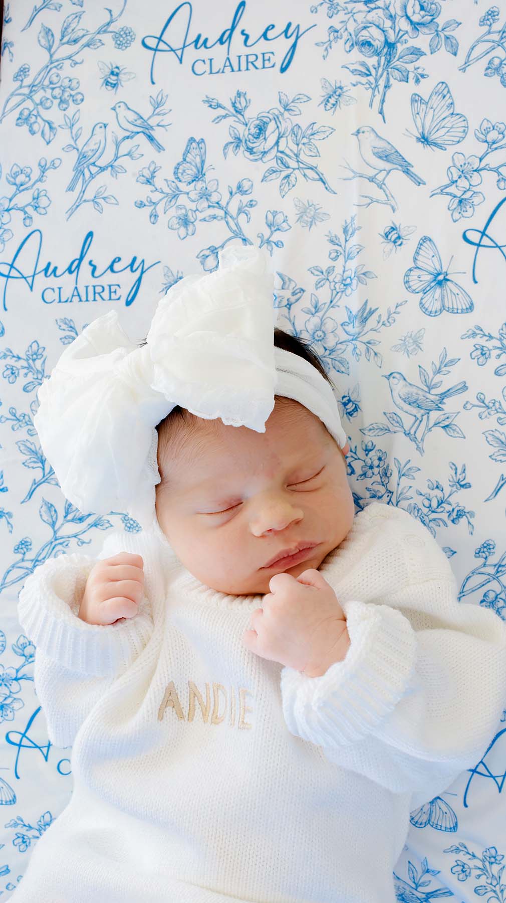Close-up of a sleeping newborn, hands clasped, on a Caden Lane blue knit swaddle in an Enchanted Meadow Toile print featuring butterflies, reading "Audrey Claire," paired with a white bow.