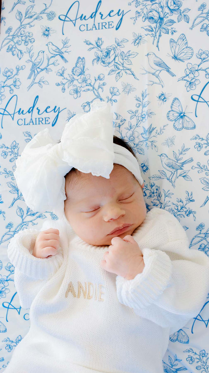 Close-up of a sleeping newborn, hands clasped, on a Caden Lane blue knit swaddle in an Enchanted Meadow Toile print featuring butterflies, reading "Audrey Claire," paired with a white bow.