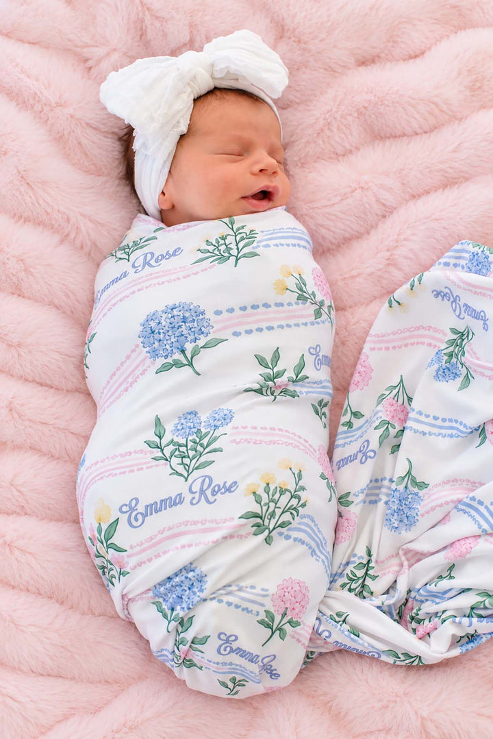 Overhead shot of a yawning newborn wearing a Caden Lane white knit swaddle in a Hydrangea Garden print featuring hydrangeas and ribbons, reading "Emma Rose," paired with a bow headband.