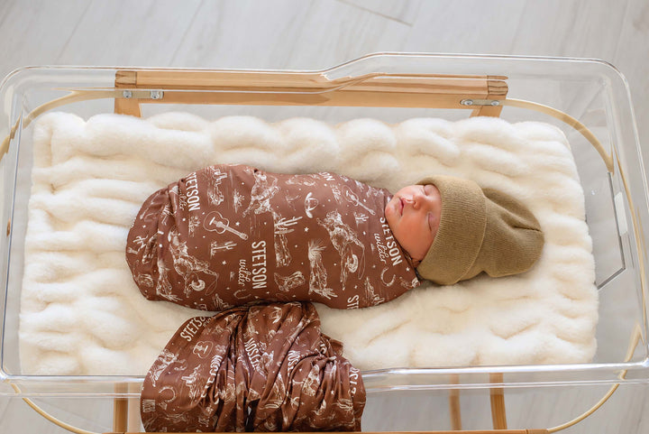 Side view of a newborn in a bassinet wearing a Caden Lane brown knit swaddle in a Wild West Toile print featuring cowboys, reading "Stetson Wilder," paired with a beanie.