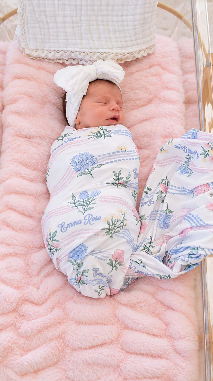 Full-length shot of a newborn in a clear bassinet, wearing a Caden Lane white knit swaddle in a Hydrangea Garden print featuring hydrangeas, reading "Emma Rose," paired with a headband.