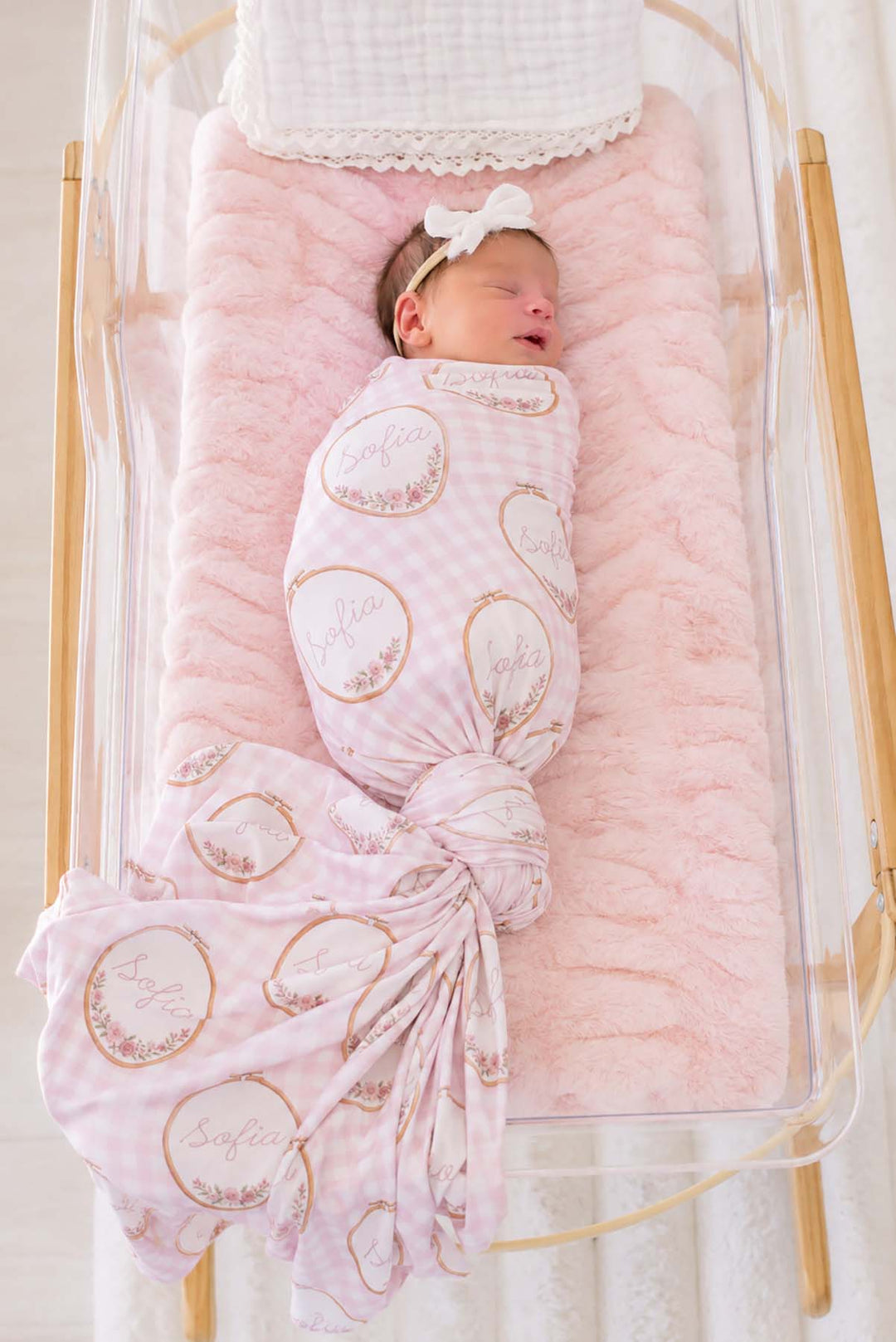 Full-length shot of a newborn, tail fanned out, wearing a Caden Lane pink knit swaddle in a Gingham Garden print featuring embroidery hoops, reading "Sofia," paired with a bow headband.