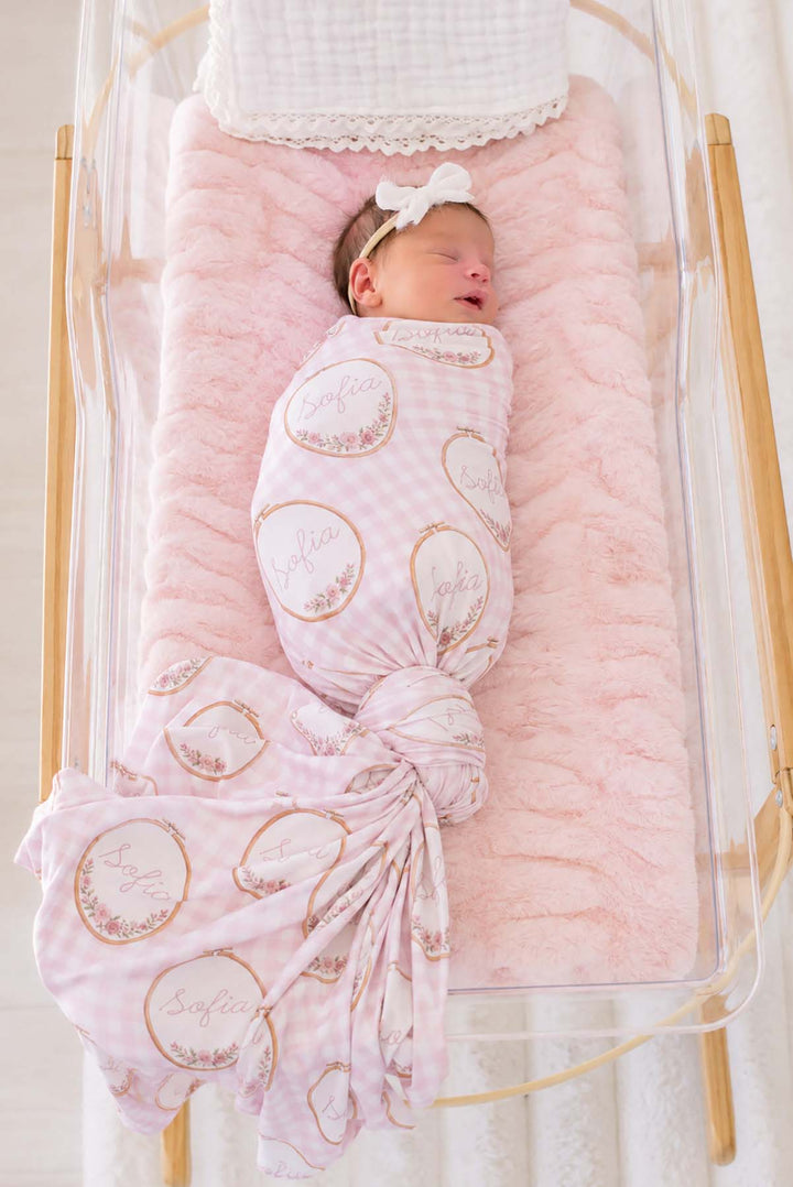 Full-length shot of a newborn, tail fanned out, wearing a Caden Lane pink knit swaddle in a Gingham Garden print featuring embroidery hoops, reading "Sofia," paired with a bow headband.