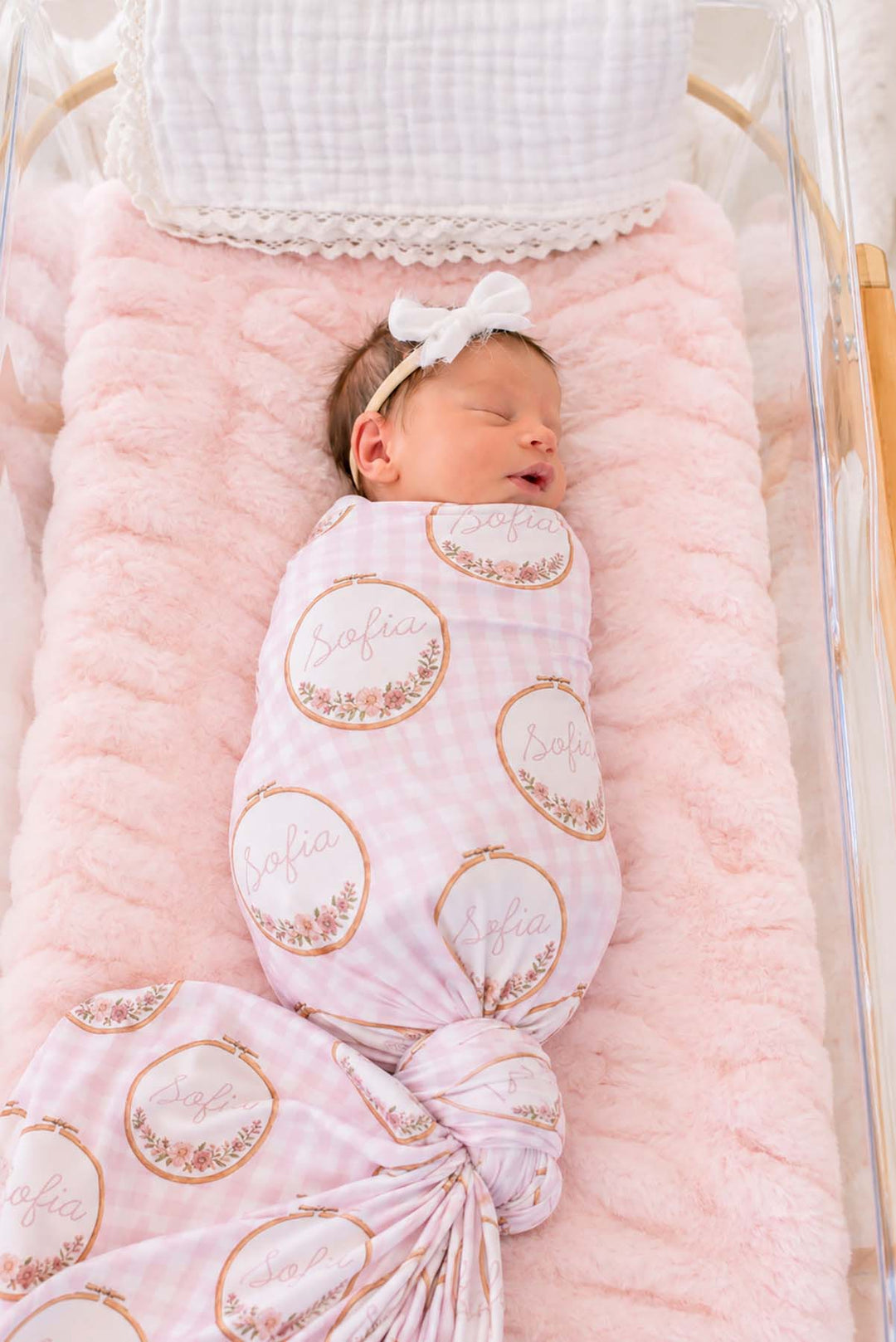 Overhead shot of a newborn in a bassinet wearing a Caden Lane pink knit swaddle in a Gingham Garden print featuring embroidery hoops, reading "Sofia," paired with a bow headband.