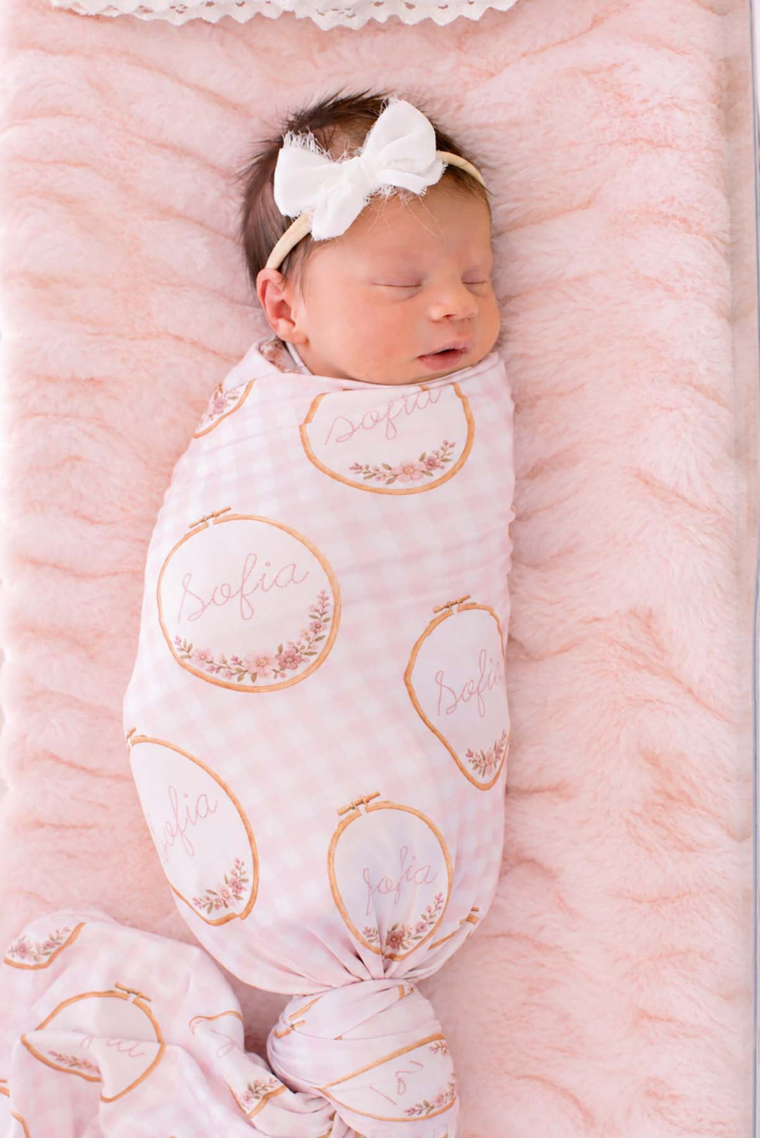 Overhead shot of a newborn wearing a Caden Lane pink knit swaddle, knotted at the feet, in a Gingham Garden print featuring embroidery hoops, reading "Sofia," paired with a bow.
