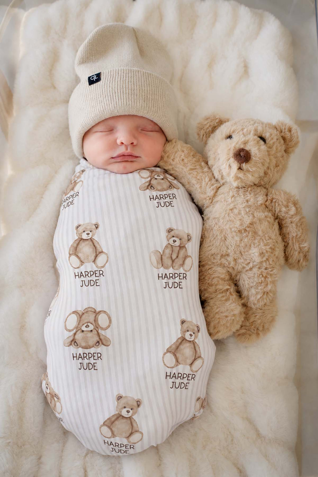 Overhead shot of a sleeping newborn wearing a Caden Lane cream knit swaddle in a Bear Stripe print featuring teddy bears, reading "Harper Jude," paired with a plush bear.