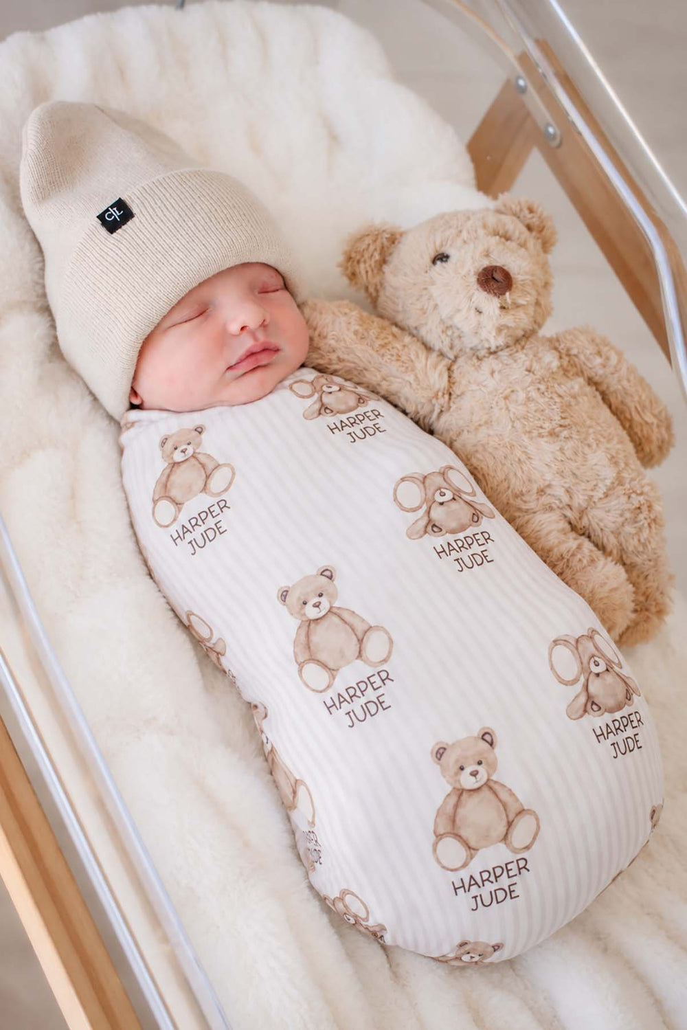 Close-up of a newborn snuggled against a teddy bear, wearing a Caden Lane cream knit swaddle in a Bear Stripe print featuring bears, reading "Harper Jude," paired with a beanie.