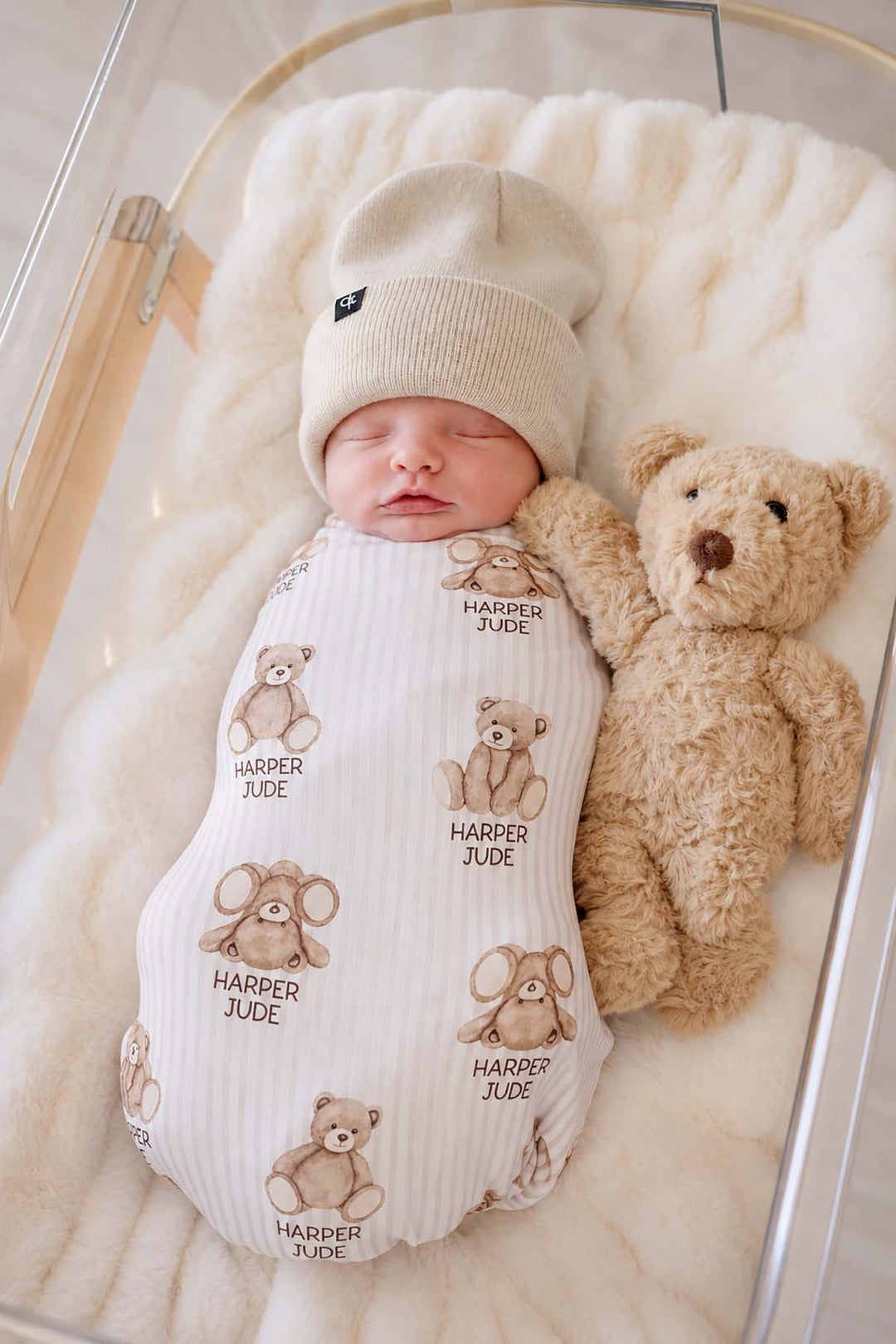 Full-length shot of a newborn in a clear bassinet wearing a Caden Lane cream knit swaddle in a Bear Stripe print featuring bears, reading "Harper Jude," paired with a teddy.
