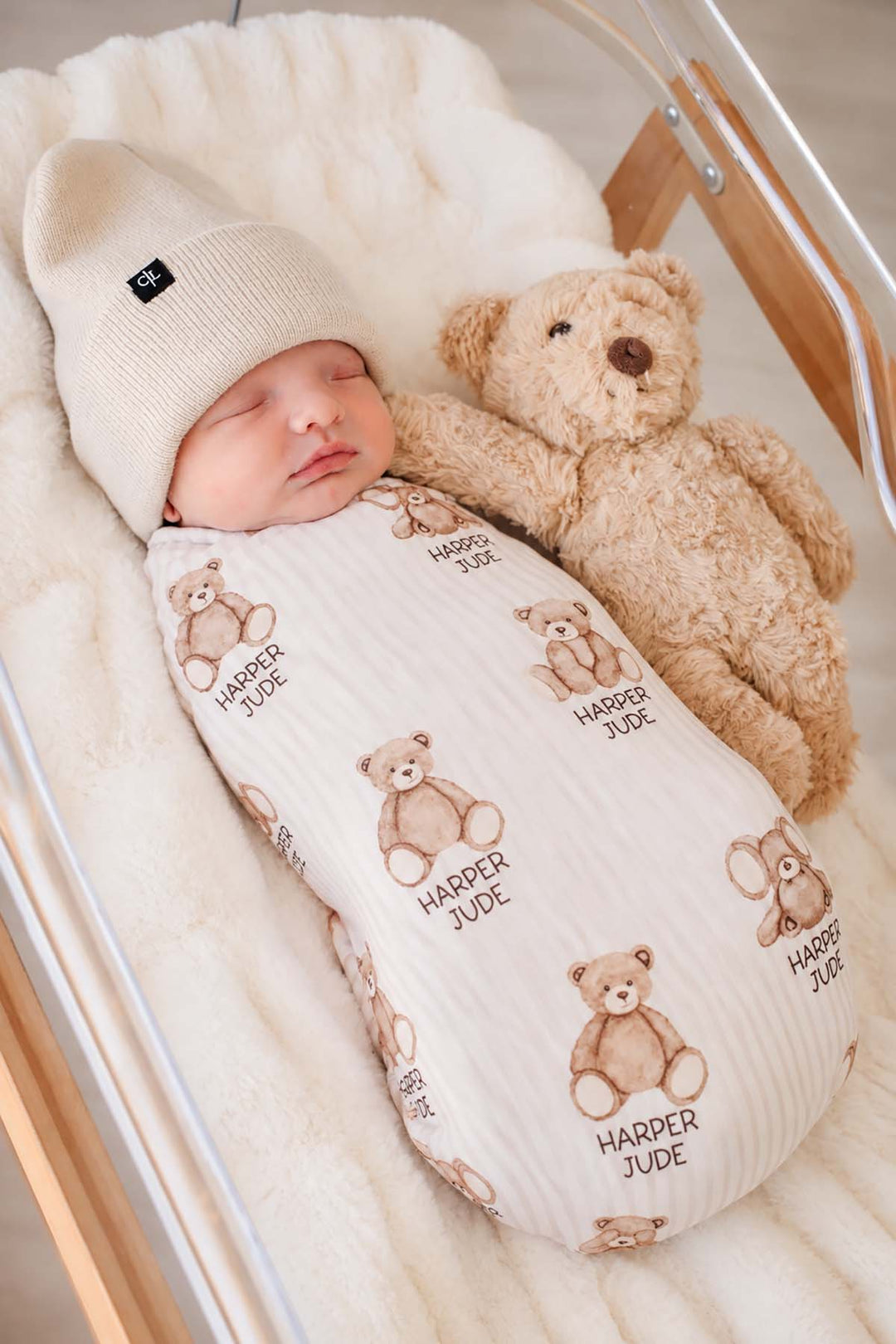Angled shot of a newborn beside a plush bear, wearing a Caden Lane cream knit swaddle in a Bear Stripe print featuring stripes, reading "Harper Jude," paired with a beanie.