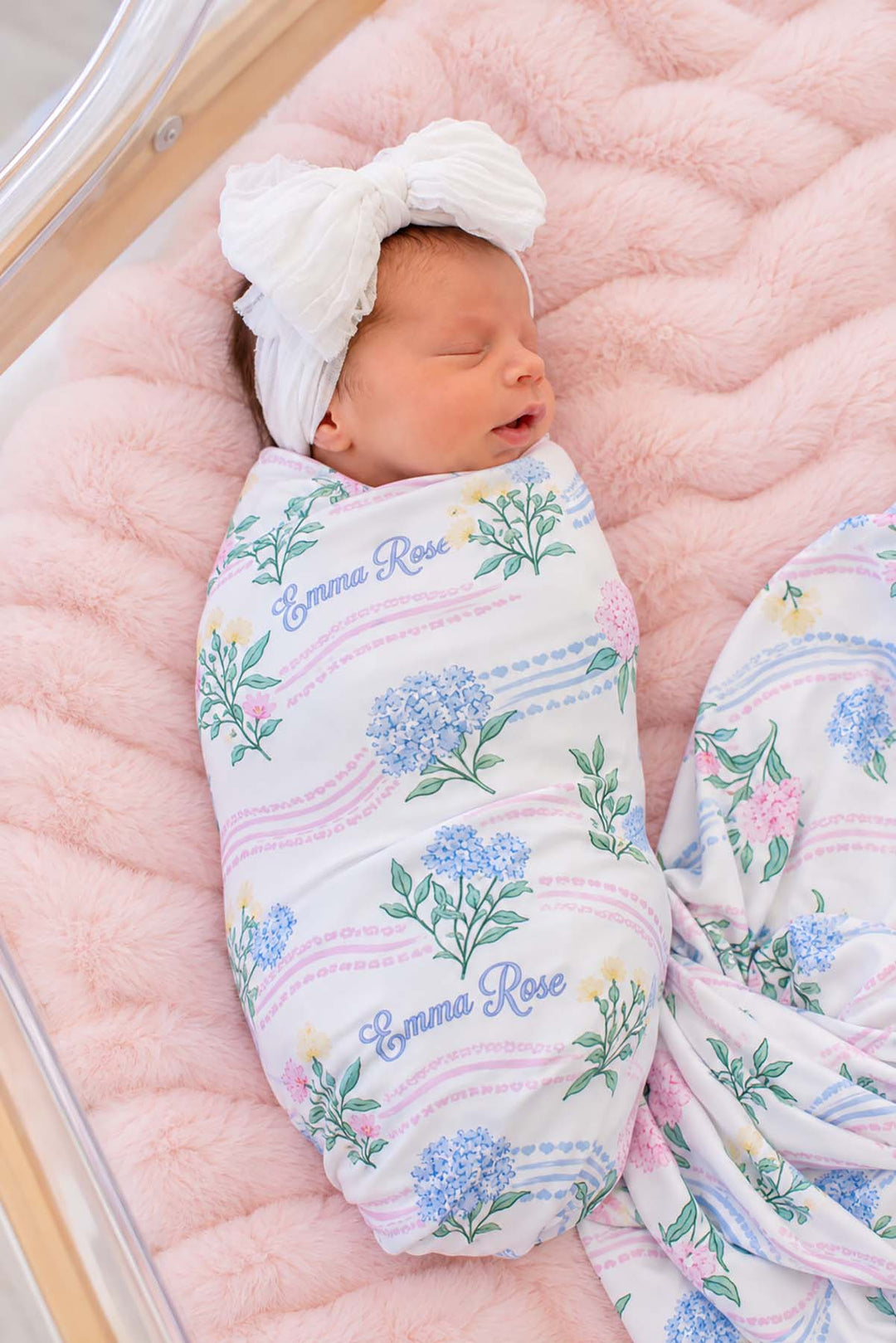 Overhead shot of a newborn in a bassinet wearing a Caden Lane white knit swaddle in a Hydrangea Garden print featuring hydrangeas, reading "Emma Rose," paired with a bow headband.