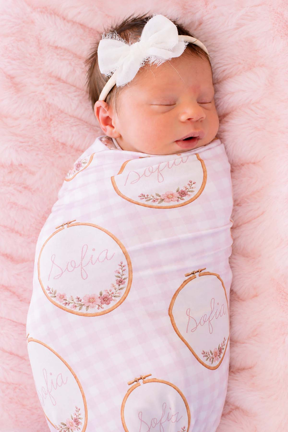 Close-up of a sleeping newborn wearing a Caden Lane pink knit swaddle in a Gingham Garden print featuring floral embroidery hoops, reading "Sofia," paired with a white tulle bow headband.