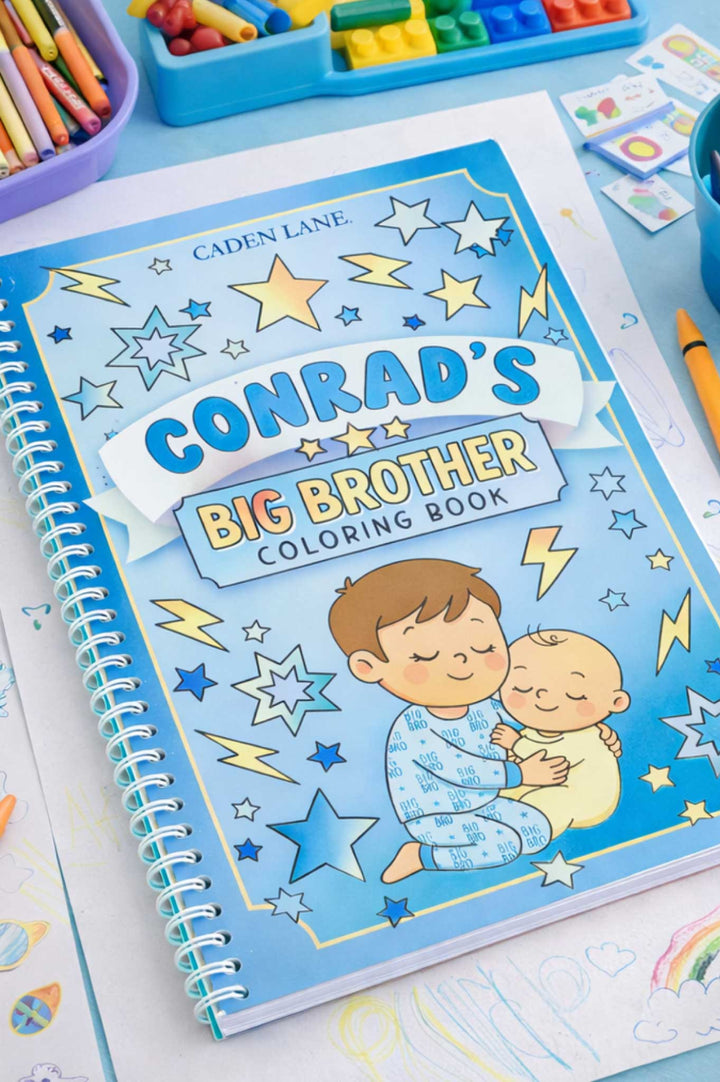 Caden Lane 'Conrad's Big Brother Coloring Book' with a blue star and lightning bolt cover featuring a big brother hugging a baby, displayed on a desk with markers, crayons, and building blocks