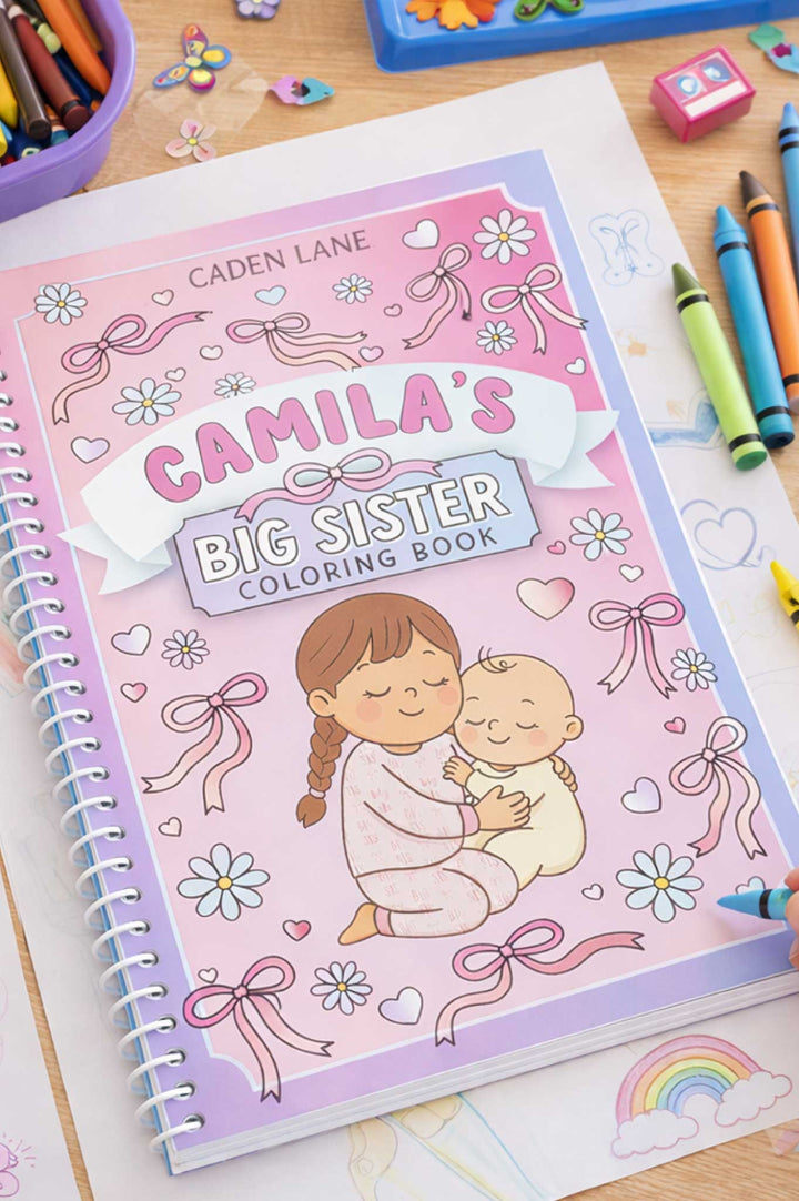 Caden Lane 'Camila's Big Sister Coloring Book' with a pink floral and bow cover featuring a big sister hugging a baby, displayed on a desk with crayons and a coloring page in progress