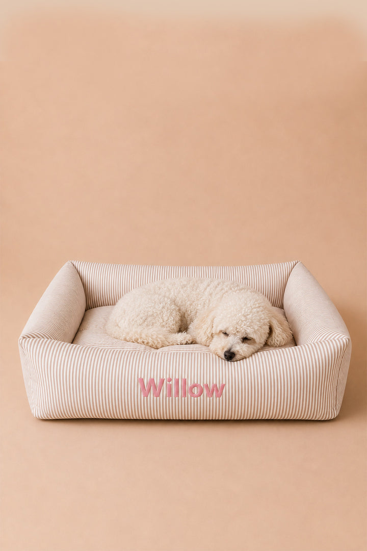 A small cream-colored poodle lies resting inside a Lane & Leash beige ribbed rectangular dog bed with pink embroidered text reading 'Willow' on the front, set against a solid warm beige background.