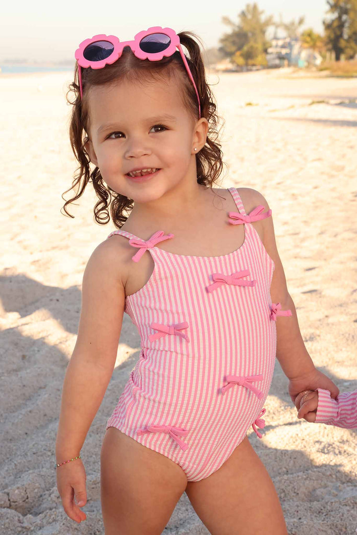 girl standing on the beach in pink bow seersucker one piece swimsuit with pink floral shaped sunglasses