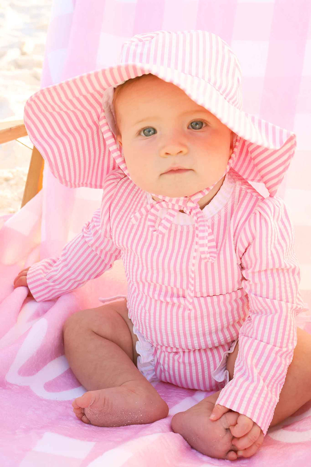 baby girl sitting on beach lounger in pink seersucker one piece swimsuit with matching hat 