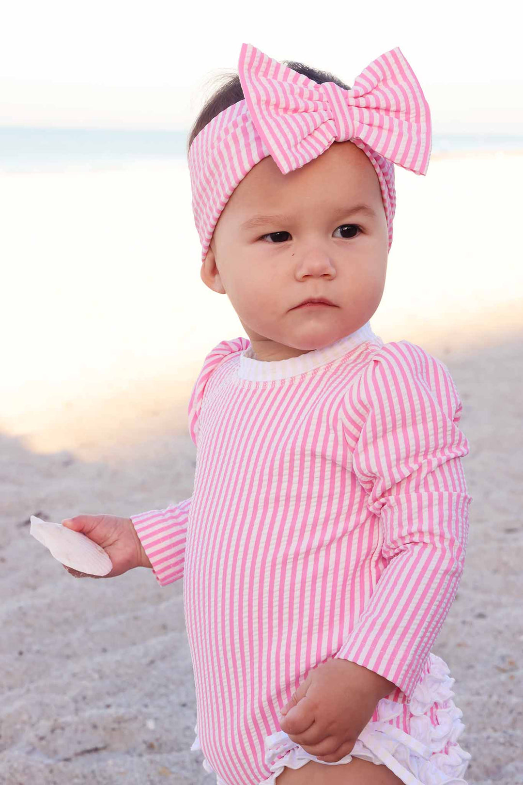 baby girl holding seashell on beach wearing pink seersucker swimsuit with matching bow