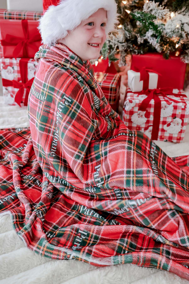 Caden Lane personalized Christmas name blanket in classic red and green plaid wrapped around a smiling child wearing a Santa hat, creating a cozy holiday scene perfect for Christmas gifts, family traditions, and festive home décor.