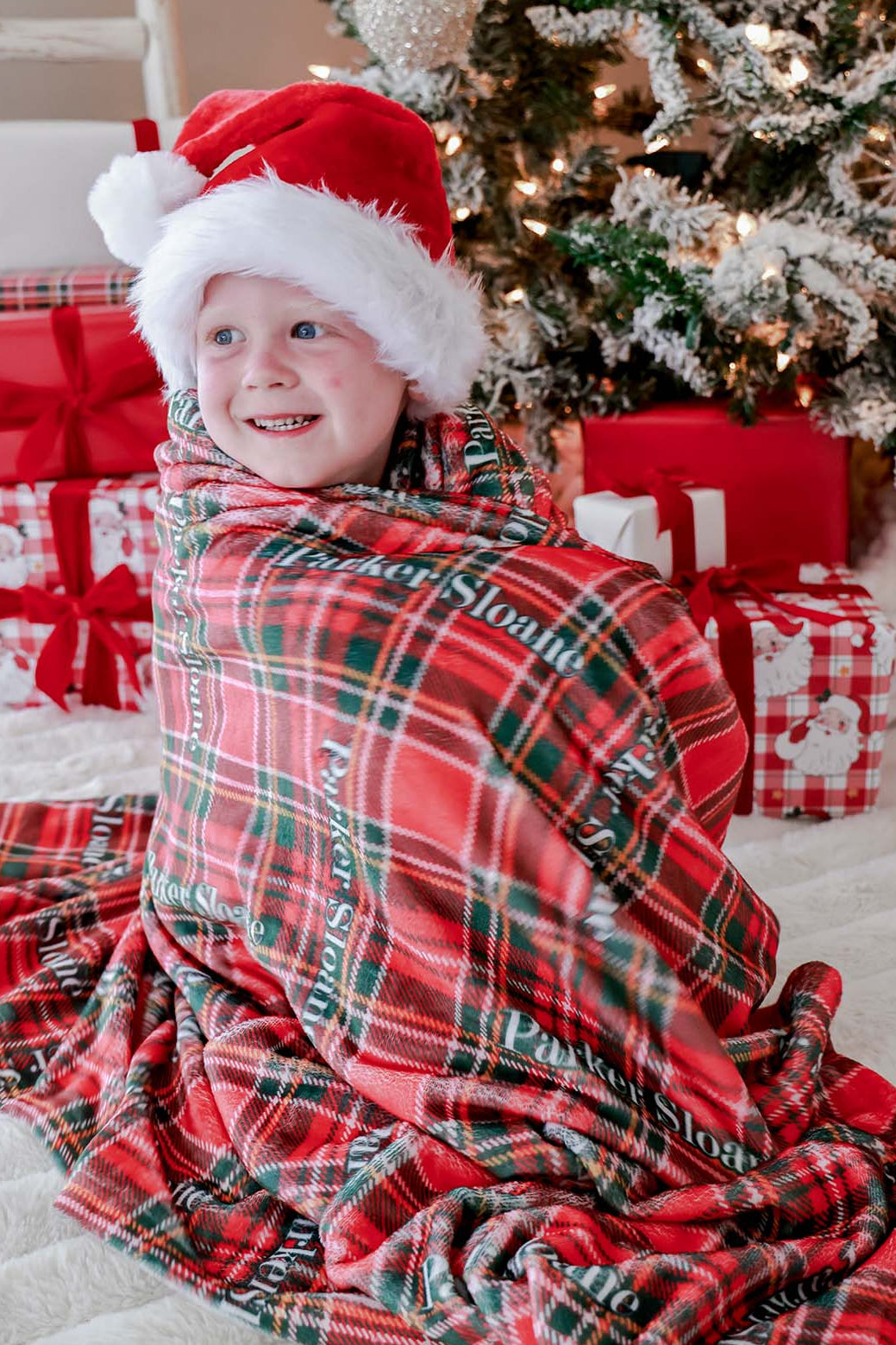 Caden Lane personalized Christmas name blanket in classic red and green plaid wrapped around a smiling child wearing a Santa hat, perfect for cozy holiday moments, matching family pajamas, and personalized Christmas gifts.