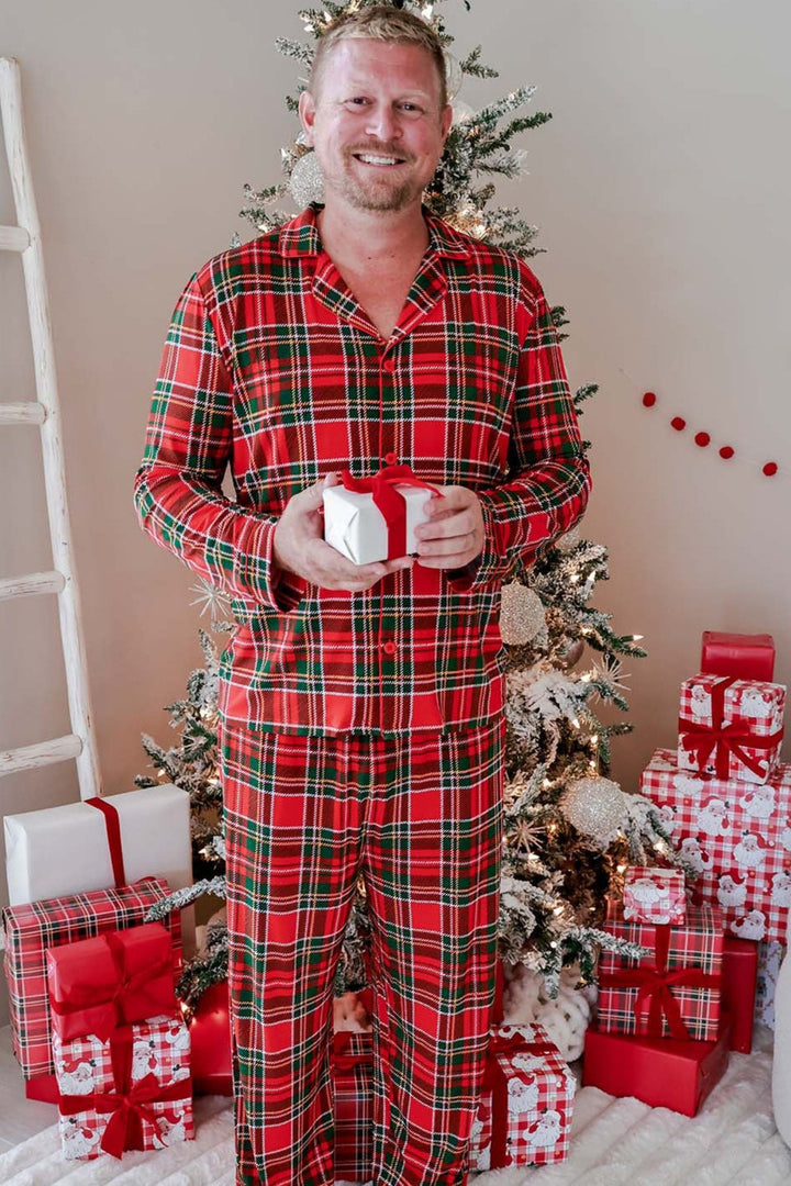 A man wearing a Caden Lane red and green plaid pajama set stands barefoot in front of a decorated Christmas tree with wrapped gifts, holding a small white gift box.