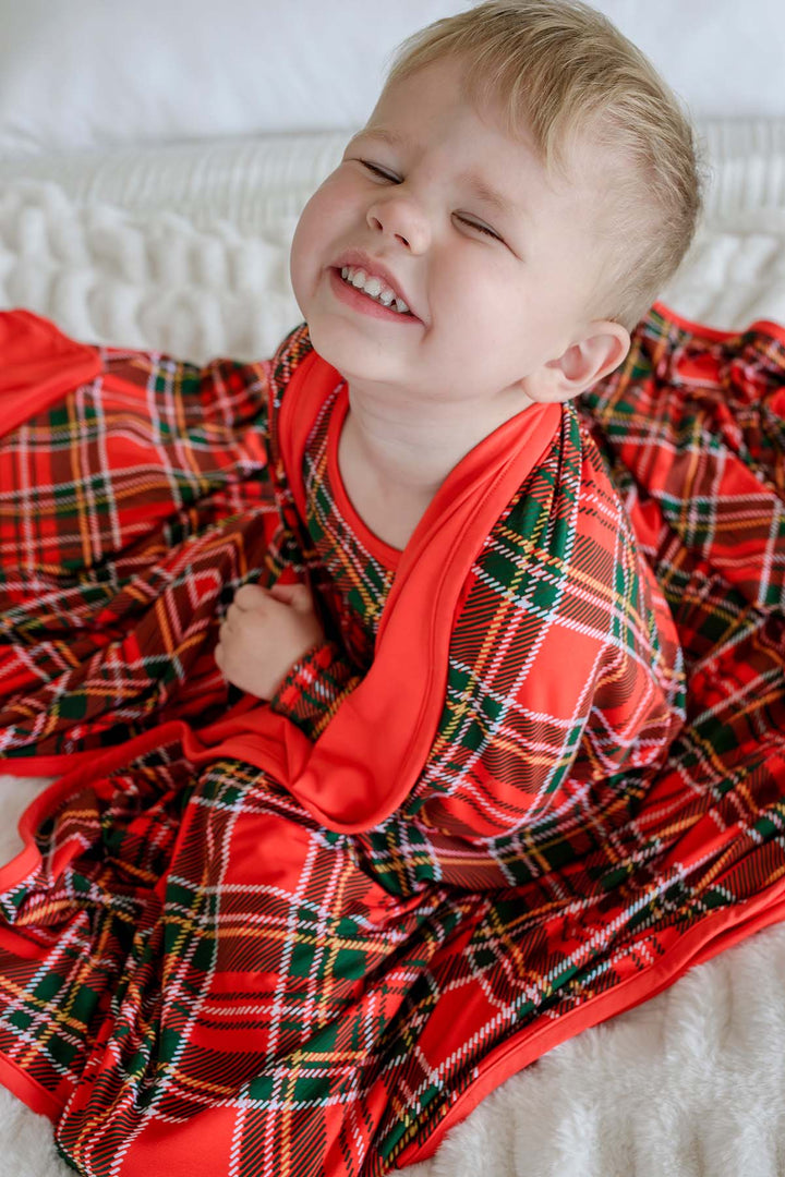 Caden Lane smiling toddler wrapped in a soft red and green plaid bamboo Christmas blanket while wearing matching holiday pajamas, perfect for cozy winter mornings, Christmas celebrations, and family memories.