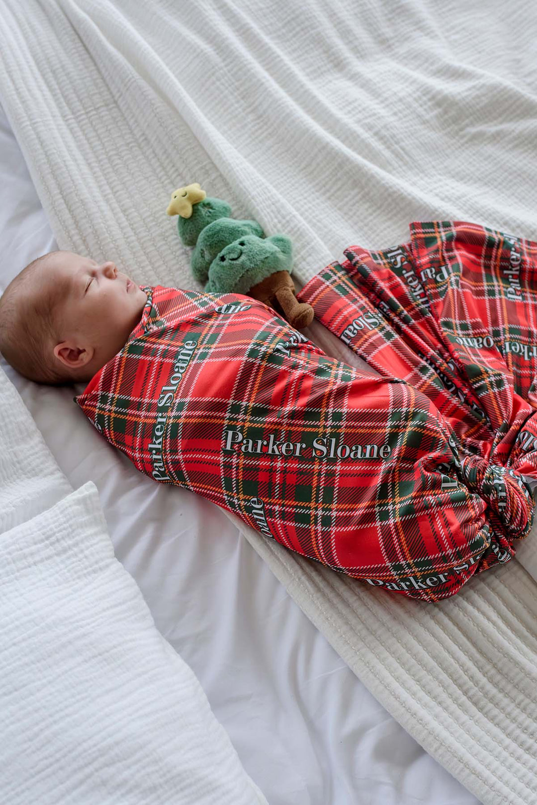 Newborn wrapped in a Caden Lane personalized plaid Christmas swaddle blanket with custom name, soft bamboo fabric, and festive red tartan print for baby's first Christmas and holiday newborn photos.