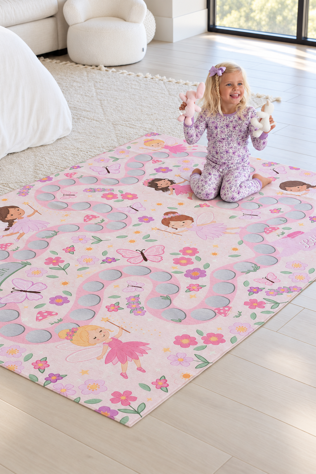 Smiling girl holding plush toys on a personalized Caden Lane fairy garden playroom rug featuring pink fairies, flowers and butterflies.