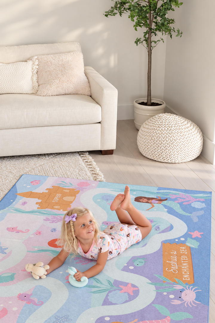 Young girl sitting on a Caden Lane mermaid playroom rug decorated with pastel mermaids, fish, starfish, coral and winding ocean paths.