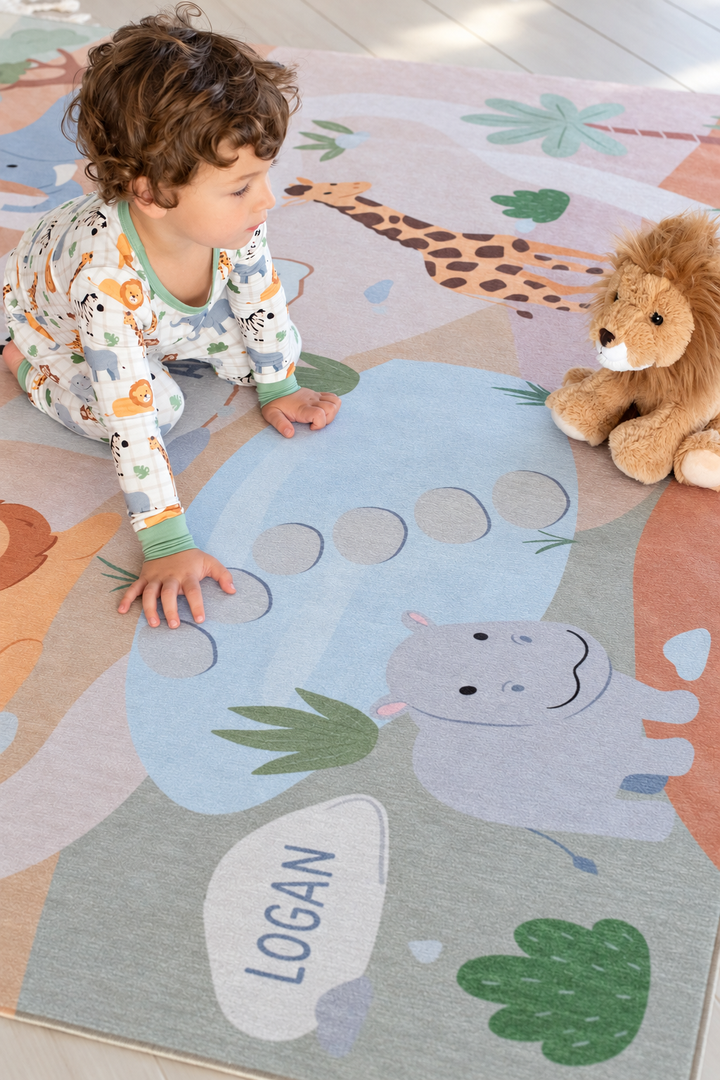 Toddler playing beside a plush lion on a personalized Caden Lane safari playroom rug featuring a giraffe, hippo and jungle scenery.