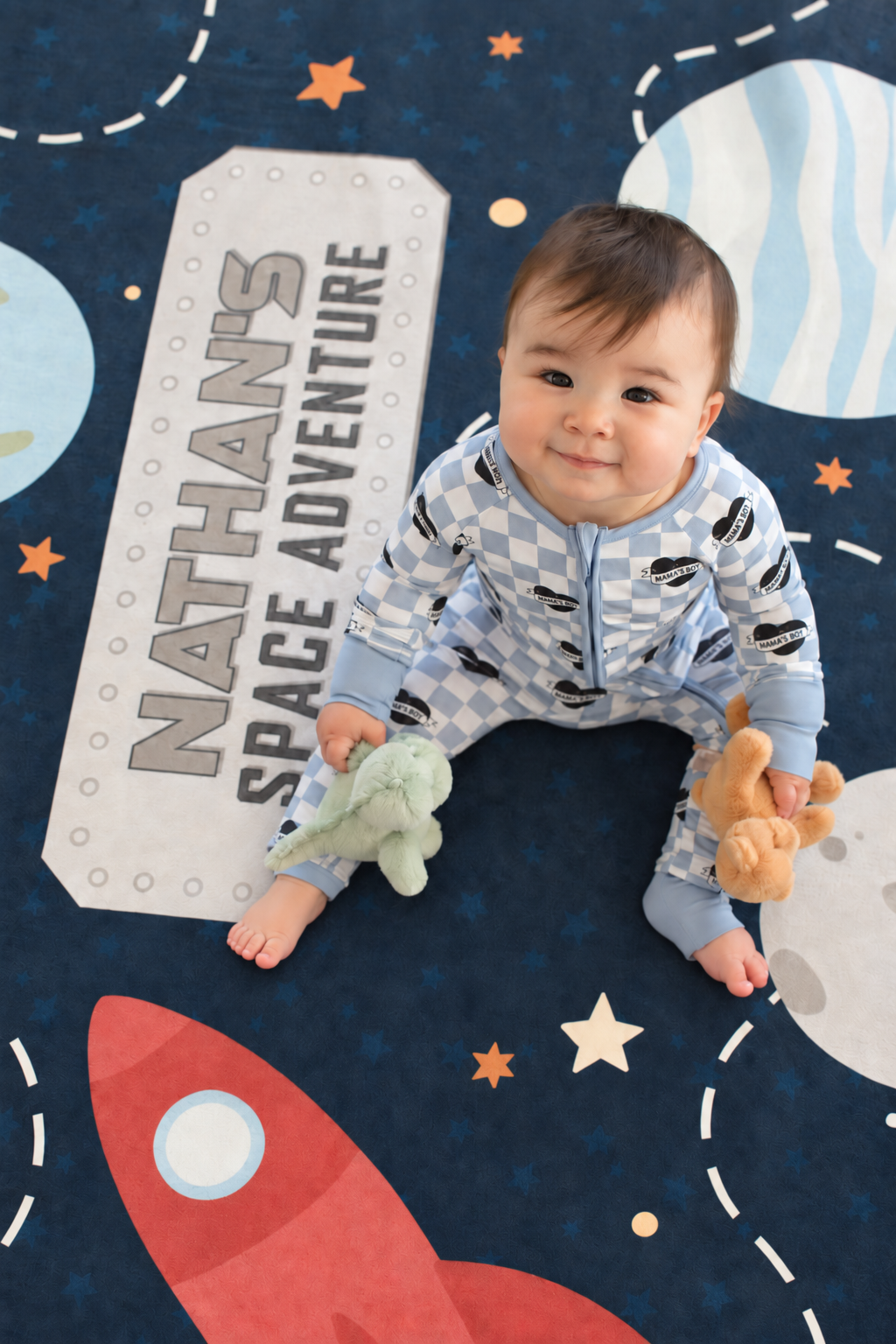 Baby holding plush toys on a personalized Caden Lane outer space playroom rug featuring colorful planets, moons, stars and rocket ships.