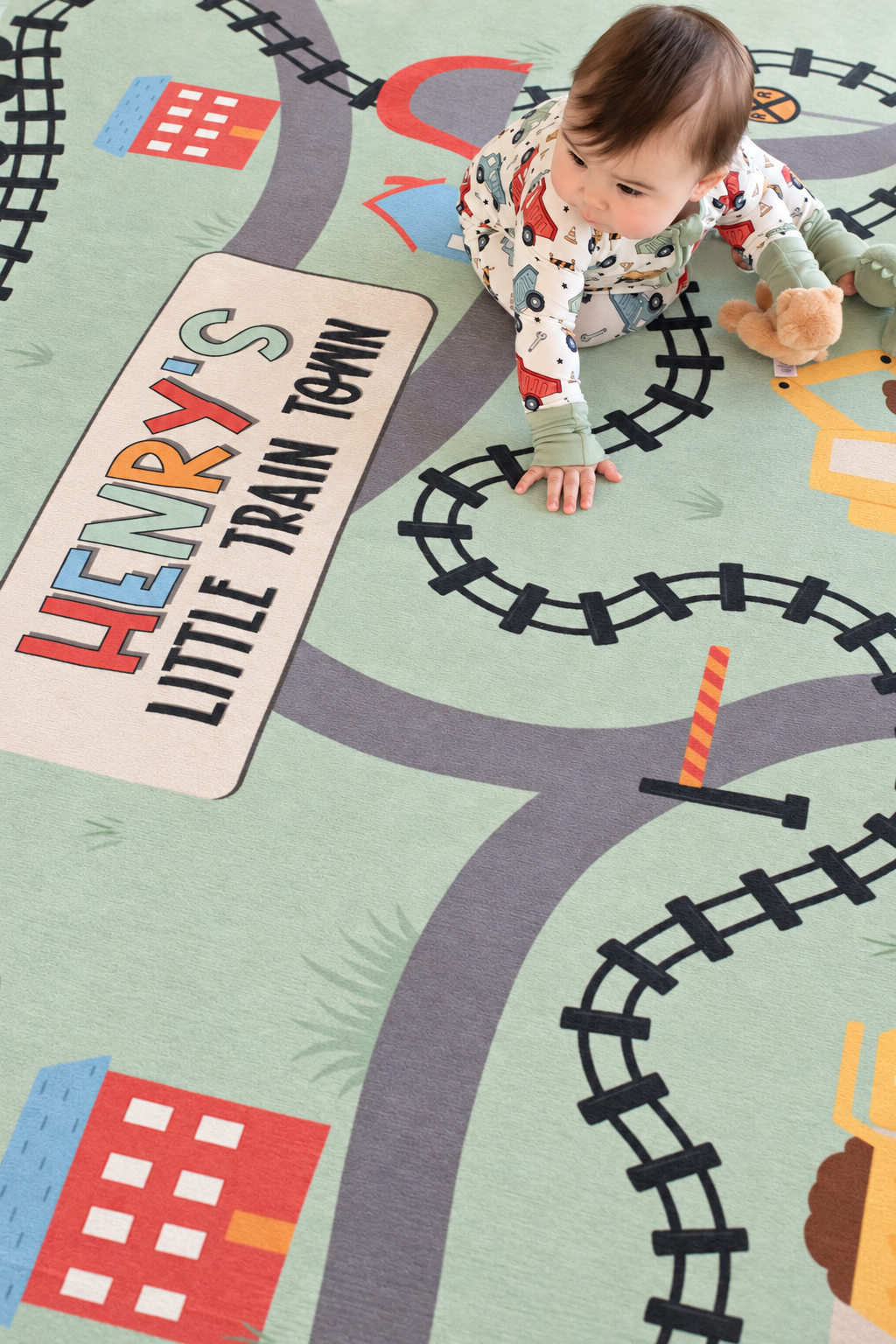Overhead view of a baby playing on a personalized Caden Lane train playroom rug featuring railroad tracks, buildings and construction equipment.