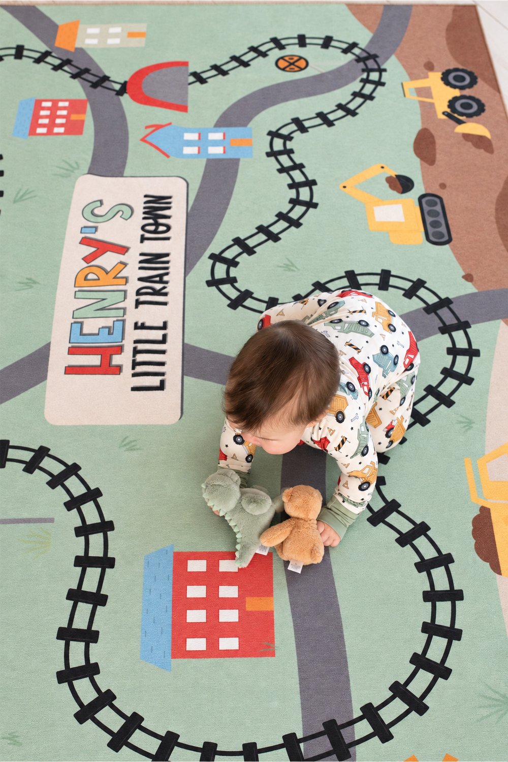 Baby playing with plush toys on a personalized Caden Lane train-themed playroom rug with colorful tracks and construction vehicles.