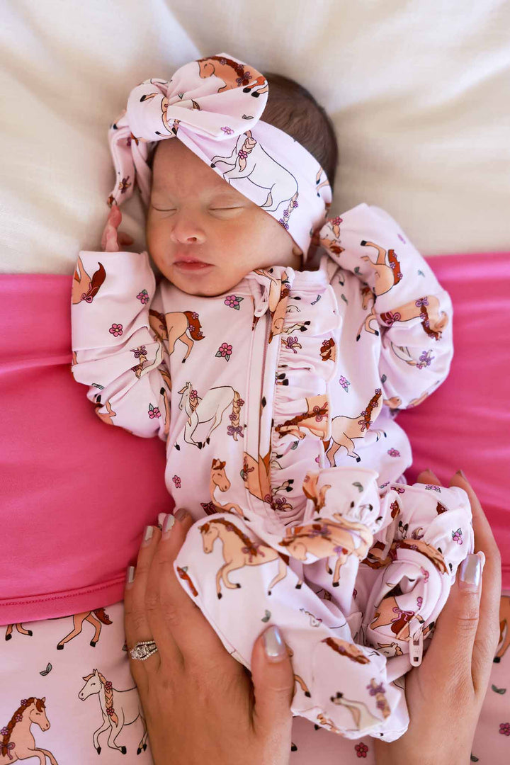 newborn girl wearing pink pony ruffle zipper footie with a matching blanket behind her