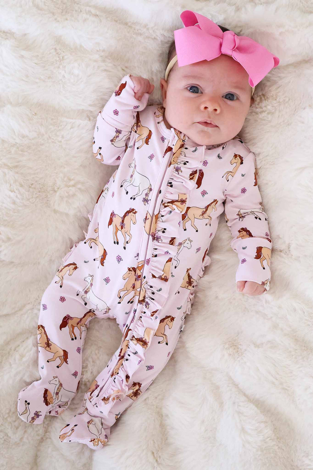 baby girl laying on fluffy white blanket wearing the pony club pink ruffle zipper footie with a matching pink ribbon bow headband 