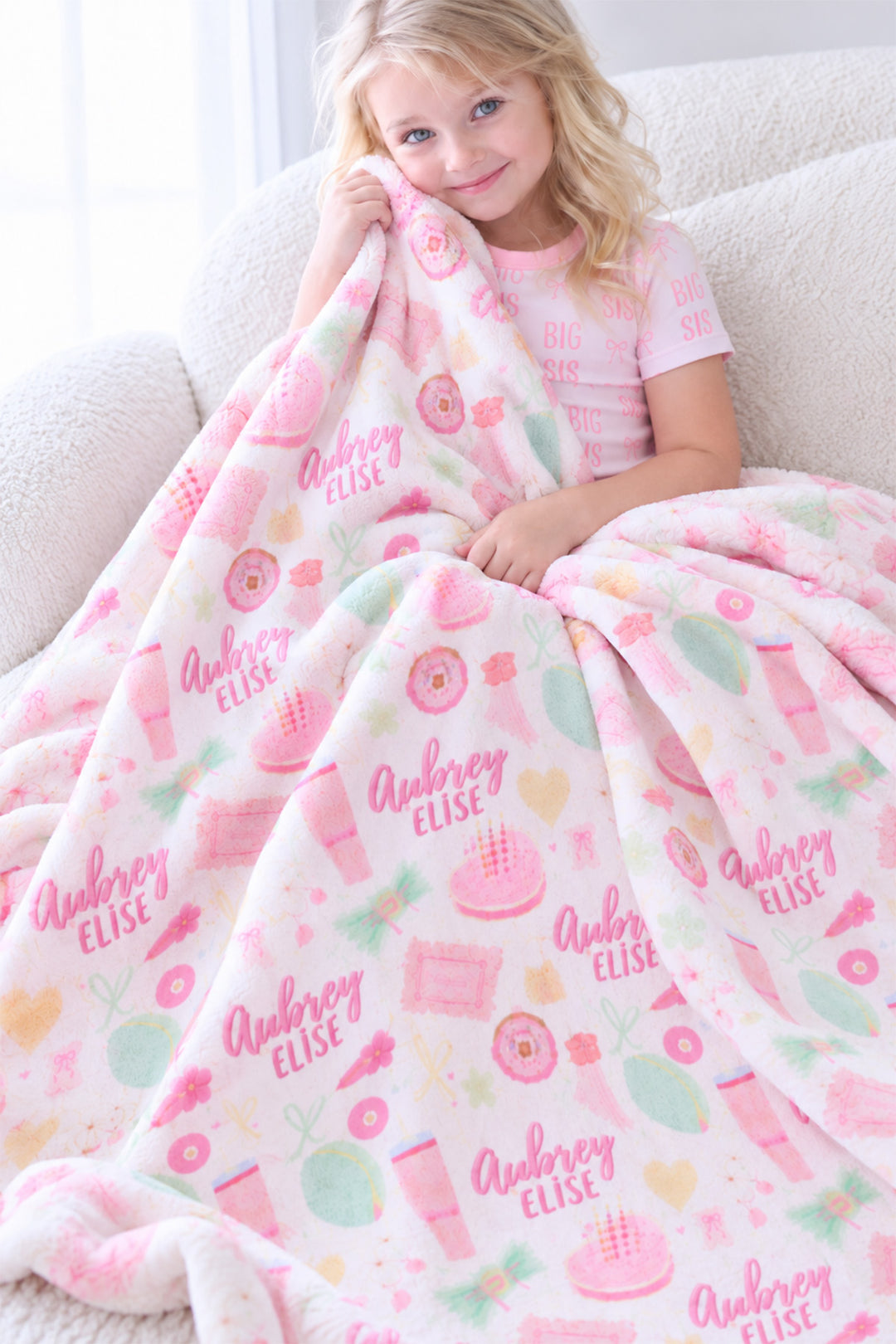 Smiling girl in "Big Sis" pajamas sitting on a couch wrapped in a personalized Caden Lane Pretty in Pink blanket, showing pink and mint icons and custom name print
