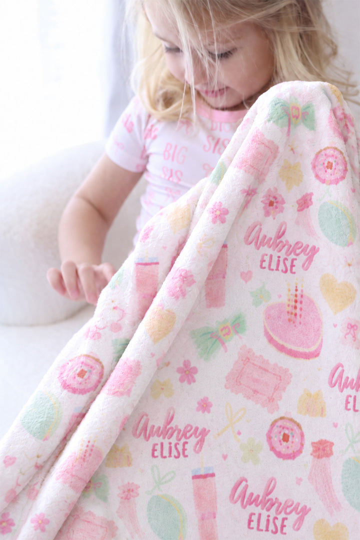 Close-up of a girl in "Big Sis" pajamas holding up a personalized Caden Lane Pretty in Pink blanket, showing birthday cake, heart, and custom name detail