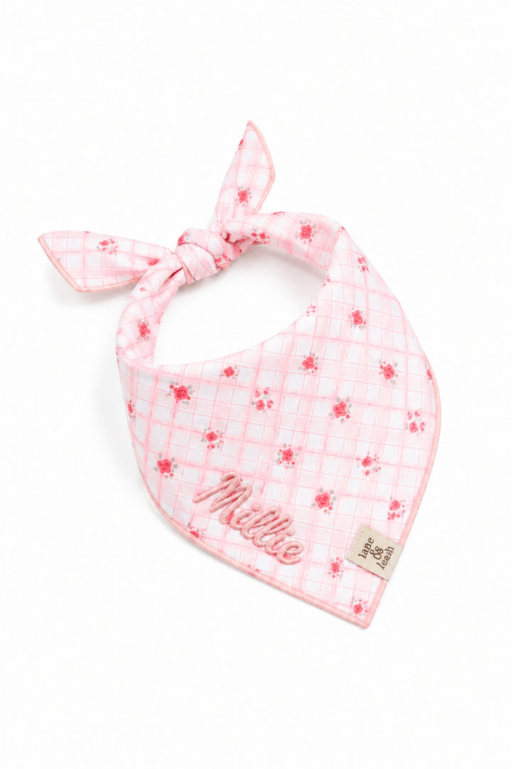 A Lane & Leash pink plaid pet bandana with small red floral motifs and the name 'Mille' embroidered on the front, tied in a knot on a white background.