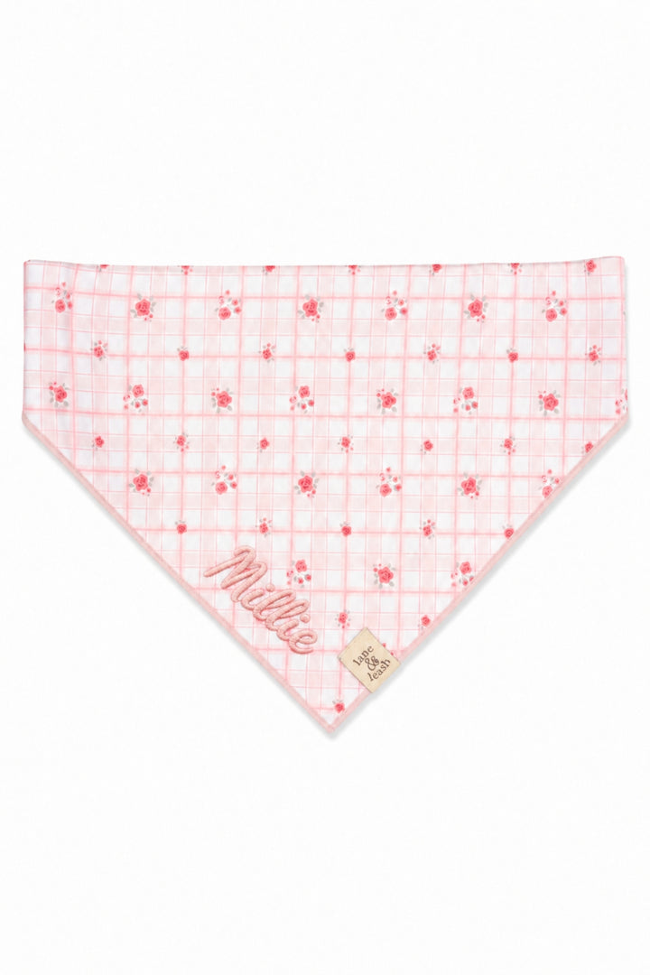 A Lane & Leash light pink pet bandana featuring a red grid pattern, small red floral motifs, and the name 'Millie' embroidered in red script on one corner, shown on a plain white background.