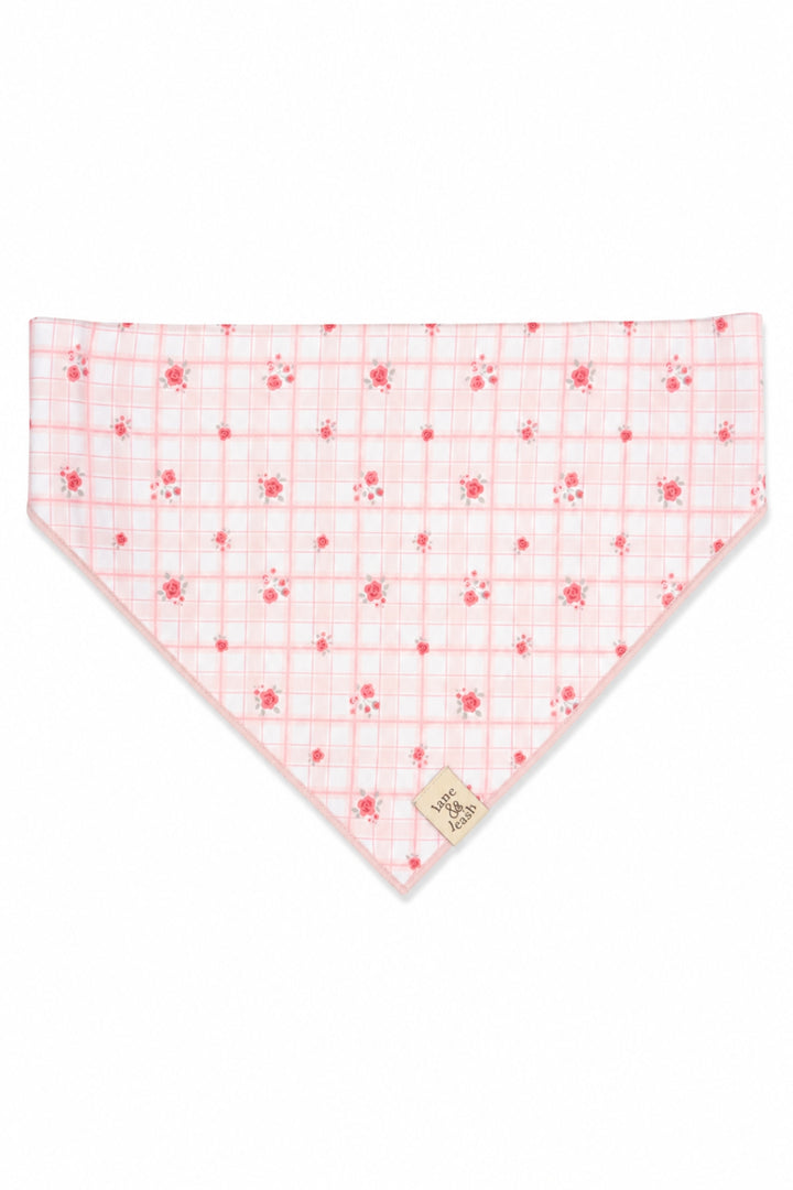 A triangular Lane & Leash bandana-style pet bib in white fabric with a light pink grid pattern and small pink floral motifs, featuring a small beige fabric brand tag near the bottom point.