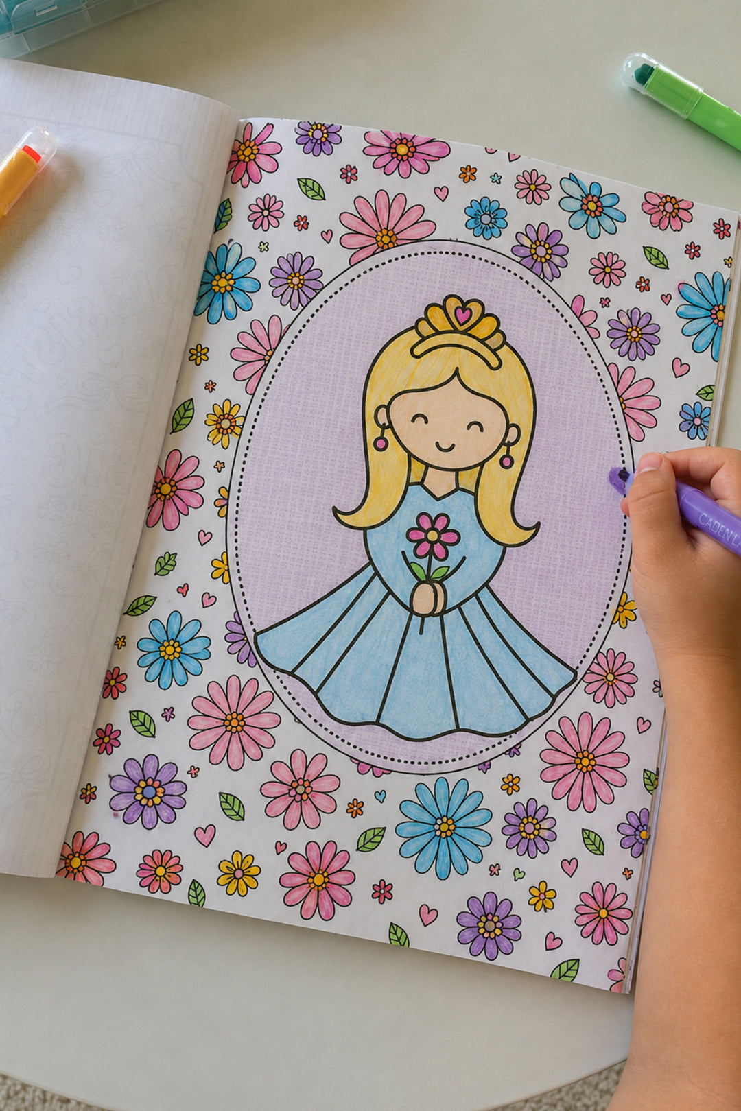 Child's hand coloring a princess page in the Caden Lane Enchanted Princess Coloring Book, using a purple Caden Lane marker surrounded by a colorful floral border