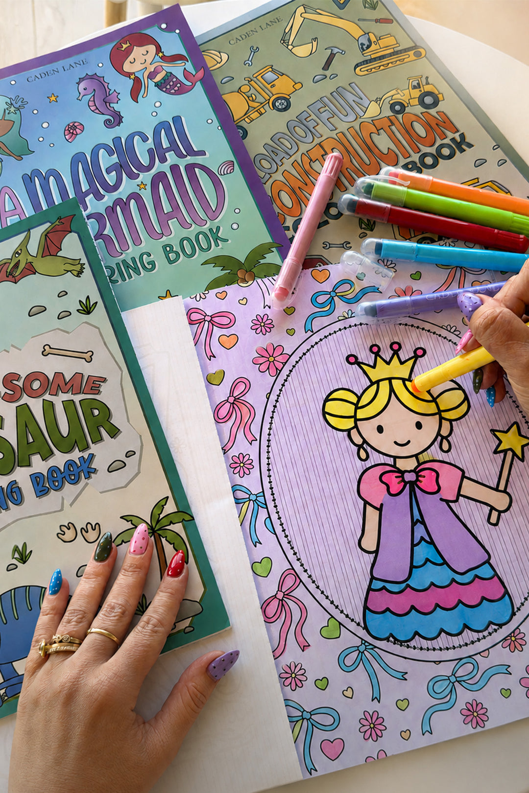Caden Lane coloring books including Enchanted Princess, Magical Mermaid, and Construction titles fanned out on a table, with a child's hand using Caden Lane gel crayons and markers to color a princess holding a wand.